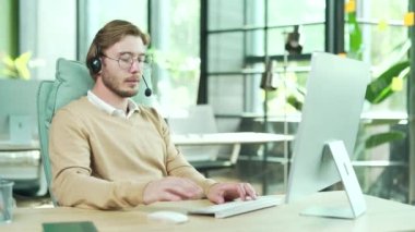 Kendine güvenen genç iş adamı destek yöneticisi kablosuz kulaklığı ile bilgisayar ekranına bakıyor ve çevrimiçi toplantı konferans video görüşmesi ile konuşuyor modern ofis içinde iş yerinde webcam kullanarak