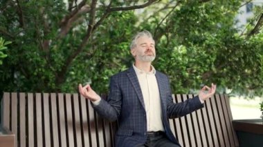 Rahat sakin işadamı, gözleri kapalı, yoğun çalışma gününden sonra yoga yapıyor modern ofis merkezinin yanındaki bankta oturuyor, dinlenen olgun bir çalışana gülümsüyor ve dışarıda huzur içinde nefes alıyor.
