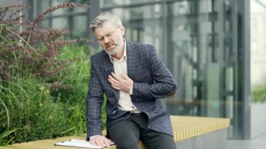 Aşırı çalışan olgun bir işadamı, modern iş merkezinin yanındaki bankta otururken kalbinde ani bir ağrı hisseder. Stresli erkek, göğsünde kalp krizi geçirerek kalp krizi geçirir.
