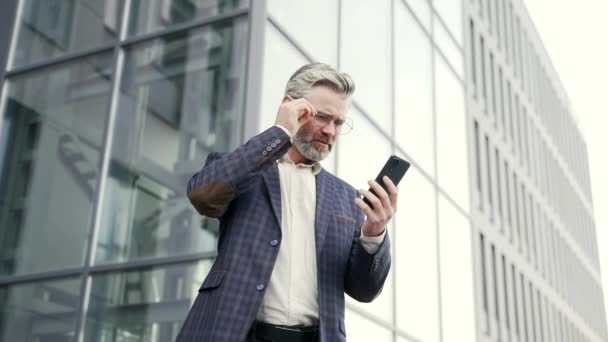 悲しい成熟したビジネスマンは 現代のビジネスセンターで携帯電話に対処する銀行の財務上の問題とのメッセージを受信しますエレガントな動揺男は スマートフォンの屋外で悪いニュースを読んで電話を見て — ストック動画