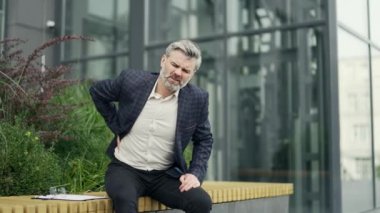 Yorgun girişimci işadamı ciddi bir sırt ağrısı çekiyor modern iş merkezinin yanındaki bankta oturuyor. Sağlıksız, hasta, sırtına kramp giren bir adam. Açık havada omurga ağrısı çekiyor.