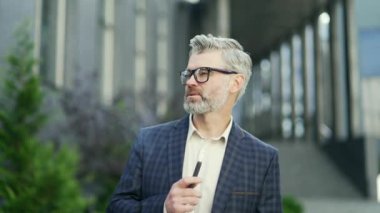 Modern iş merkezinde elektronik sigarayla buharlaşan gülümseyen yetişkin bir işadamı. Klasik takım elbiseli başarılı bir adam ofis binalarının yakınında e-sigara içiyor.