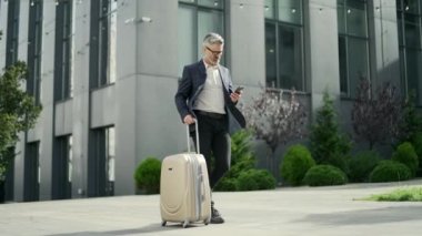 Kendine güvenen, olgun bir iş adamı, iş seyahati sırasında akıllı telefonları karıştırır. Modern ofis merkezinde resmi bir yolculuk. Bavullarını açık havada taşıyan başarılı bir adam.
