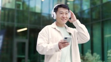 Kulaklıklı mutlu Asyalı bir adamın portresi. Müzik dinliyor ve modern iş merkezinde tek başına yürüyor. Akıllı telefonlu güler yüzlü, haftasonu açık havada eğlenen şık bir adam.