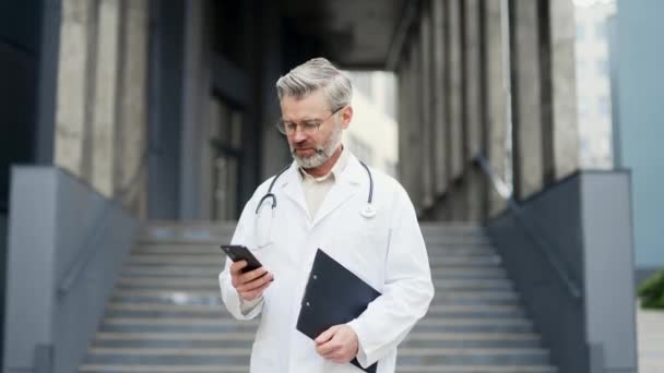 Uśmiechnięty Dojrzały Lekarz Chirurg Białej Szacie Stetoskopem Przytrzymaj Smartfon Nowoczesnych — Wideo stockowe