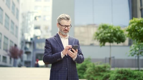 自信を持って成熟したビジネスマン起業家は 現代のビジネスセンターでスクロールブラウジングテキストメッセージを保持していますオフィスビルの近くの電話で古典的なスーツウォークでメガネを持つ成功したシニア男性 — ストック動画