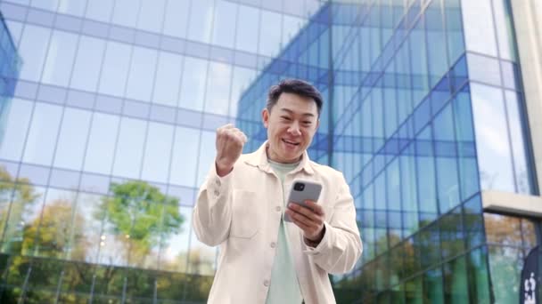 在户外的手机屏幕上读到消息后 非常快乐的亚洲男人兴奋地看着屏幕 高兴地在市中心的智能手机上看到了好消息 — 图库视频影像