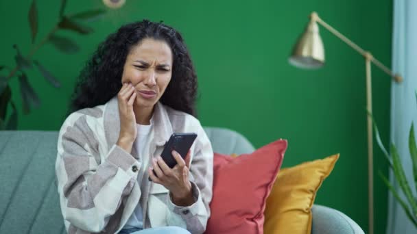 年轻的黑发卷曲女人 牙齿酸痛 牙齿酸痛 在家用智能手机单独使用 悲伤的女人 因为龋牙病或敏感的牙齿在室内而感到不适 — 图库视频影像