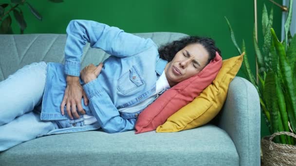 胃の痛みに苦しんでいる病気の若いヒスパニック系の女性は 自宅のリビングルームで腹感胃炎や月経痛を保持ソファの上に横たわっています婦人科の問題痛みを伴う期間概念 — ストック動画