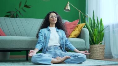 Sakin, gözleri kapalı bir kadın Lotus pozisyonunda oturmuş meditasyon yapıyor ve yavaşça nefes alıyor. Bu sırada yoga asanalarını oturma odasında dingin, kıvırcık İspanyol kadın evde tek başına meditasyon yapıyor.