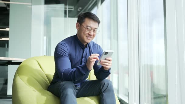 アジアのビジネスマンItプログラマーは 豆の袋の椅子に座っているスマートフォンのスクロールを保持し 現代のオフィスの職場でソーシャルメディアを見て笑顔従業員が休憩室で携帯アプリのタイプブラウジングを使用して — ストック動画