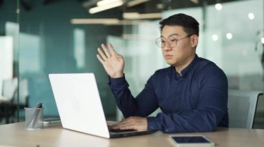 Gülümseyen Asyalı iş adamı destek müdürü gözlüklü ofis çalışanı bilgisayar ekranına bakıyor ve çevrimiçi toplantı konferans görüntüleriyle konuşuyor modern iş yerindeki webcam kullanarak. 