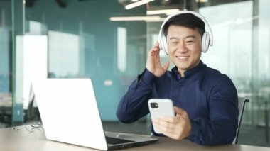 Mutlu Asyalı iş adamı kulaklıklı bilişim programcısı akıllı telefonu tut ve camdan ofiste dans eden müziği dinle gülümseyen neşeli girişimci modern işyerinde rahatlıyor ve eğleniyor. 