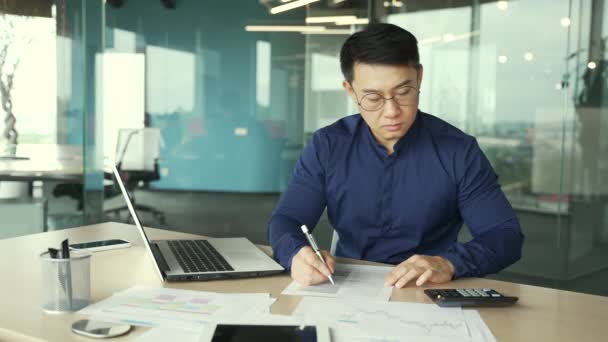現代ガラスオフィスのデスクで書類を扱う忙しいアジアの会計士事務所屋内の職場での文書やグラフで計算機に関する財務報告を数える集中ブックキーパー — ストック動画