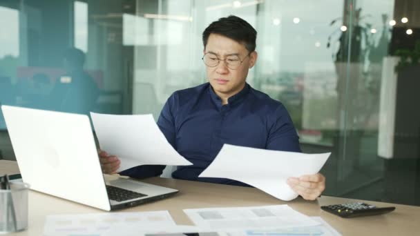 現代オフィスでの紙の作業文書に問題を抱えている神経質なアジアのビジネスマンItプログラマー開発会社のデータを分析する統計レポートで疲れを感じるストレスのある男 — ストック動画