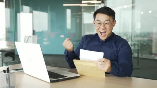 快乐的亚洲商人It程序员带着眼镜收到一封开封的信 信中写着在项目工作或加薪方面取得的巨大成功 兴奋的企业家在室内庆祝胜利 — 图库视频影像