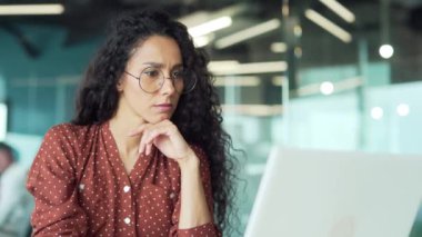 Genç, ciddi, İspanyol iş kadını masa başında oturmuş modern şirket ofis teknolojileri konseptinde dizüstü bilgisayara bakıyor. Düşünceli serbest çalışan portresi.
