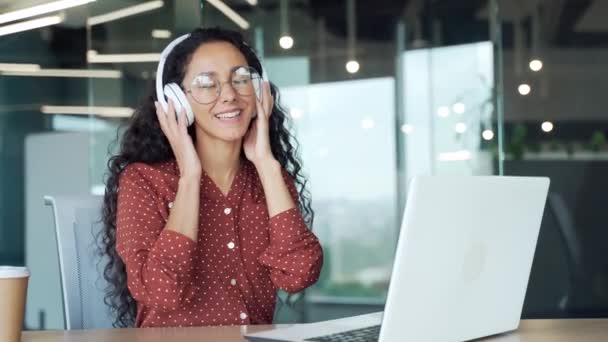 ブルネットの若い女性従業員はリラックスした音がノートパソコンで働く現代のオフィスでヘッドフォンで穏やかな音楽やラジオを楽しむリラックスした音に耳を傾ける職場の屋内で快適な学生労働者 — ストック動画