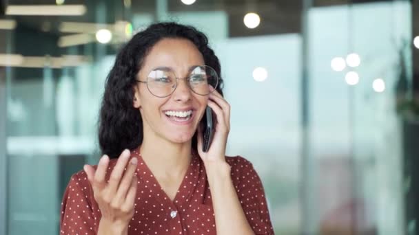 幸せな魅力的なビジネス女性従業員上の話を携帯電話に座っている現代的なオフィスにコンピュータデスクでザ 職場で屋内巻き黒髪ブルネット女性は会話笑顔を話す — ストック動画