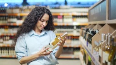 Genç ve çekici İspanyol kadın, markette alkol reyonundan şarap şişesini seçiyor. Dişi, alkol seçimini alkol bölümündeki müşterinin raflarından yapar.