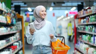 Tesettürlü genç Müslüman kadın, akıllı telefon kullanarak alışveriş listesini kontrol ediyor, süpermarketteki marketten kadın alışveriş yapıyor, hipermarkette veya gıda pazarında cep telefonu kullanıyor.
