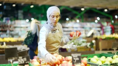 Süpermarkette, manavda ya da küçük bir dükkan sahibinde çalışan tesetteki genç bayan çalışan meyve reyonunun yanındaki elma toplayan Müslüman kadın.