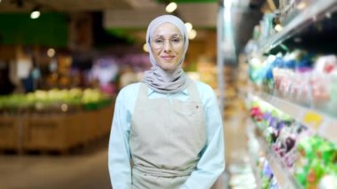 Süpermarkette tesettürlü Müslüman kadın portresi ya da önlüklü şarküteri kamera kadın satış müdürü küçük işyeri çalışanı gıda pazarında hipermarket dostu kollar çapraz