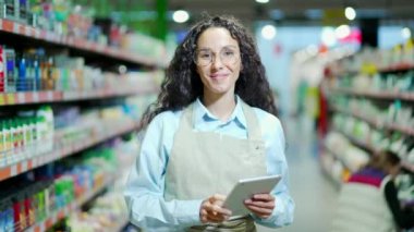 Oldukça arkadaş canlısı bir satış elemanının portresi kameraya bakar ve gülümser kadın satış asistanı ya da bir mağazanın müdürü ya da bir tabletle süpermarketin müdürü. Markette önlüklü kıvırcık esmer işçi kadrosu