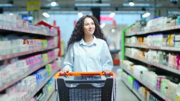 ショッピングカートを持つ幸せな若いブルネットの女性はスーパーマーケットで買い物をしていて トロリーを押して行の間で家庭用化学物質を購入して選択するつもりです — ストック動画