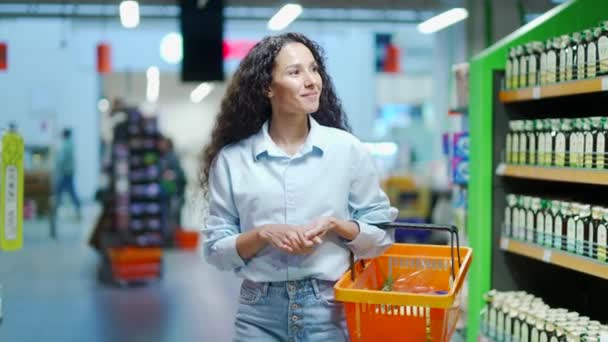 バスケットと幸せな若いブルネットの女性は 行の間に小売家庭用化学物質を購入し 選択しようとするスーパーマーケットで歩いて買い物をしています — ストック動画