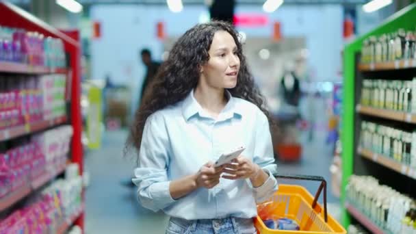 若いかなりブルネットの女性買い手顧客チェックショッピングリストを使用してスマートフォン スーパーマーケットで食料品店で購入女性買い物客ピック使用携帯電話でハイパーマーケットや食品市場 — ストック動画
