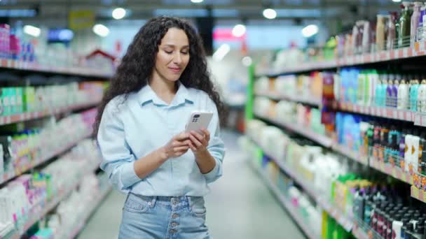幸せな若いヒスパニック系女性は列の間に立っている間 スーパーマーケットで携帯電話を使用します 屋内女性ブルネットテキストメッセージを入力し 食料品店の食品市場でスマートフォンで閲覧チャット — ストック動画