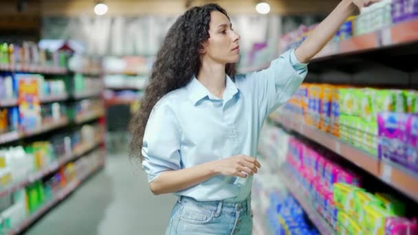 若いヒスパニックの女性は スーパーマーケットでパーソナルケア製品を選択したり 衛生のためのパッドの品揃えを格納します 女性は家庭用化学物質を選ぶ 選択を躊躇し商品の買い物を調べ — ストック動画