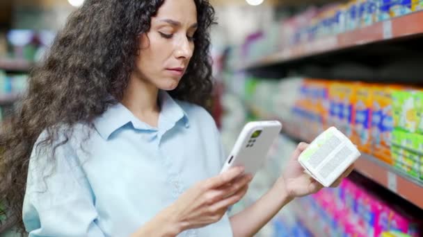 女性の顧客は スーパーマーケットで携帯電話で製品情報をチェック女性のショッピング店で商品を選択スマートフォンインターネット技術を使用して買い物客は オンライン小売割引ハイパーマーケットを購入 — ストック動画