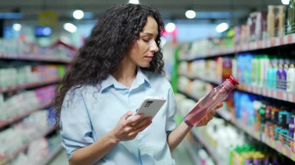 女性の顧客は スーパーマーケットで携帯電話で製品情報をチェック女性のショッピング店で商品を選択スマートフォンインターネット技術を使用して買い物客は オンライン小売割引ハイパーマーケットを購入 — ストック動画