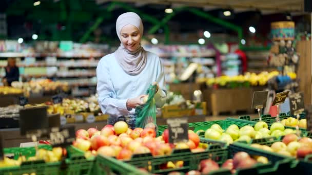 ヒジャーブ州の若いイスラム教徒の女性は スーパーマーケットや市場の食料品店の棚に果物を選択し ピックアップし エコバッグに入れて女性の顧客は ハイパーマーケットで再利用可能なパッケージにリンゴをスロー — ストック動画