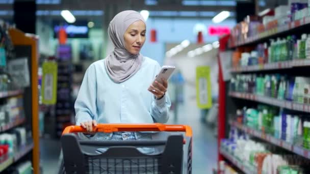 ヒジャーブ州の若いイスラム教徒の女性スマートフォンを使って買い物リストをチェックバイヤーの顧客 スーパーマーケットで食料品店で購入女性の買い物客は ハイパーマーケットや食品市場で使用携帯電話を選ぶ — ストック動画