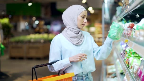 スーパーで買い物をするヒジャーブの若いイスラム教徒女性スーパーで商品を選ぶ買い物をする棚の間に籠を持つ幸せな女性の顧客商品セクションを通して閲覧 — ストック動画