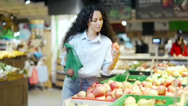 年轻漂亮的黑发女人在杂货店超市或市场的货架上挑选和采摘水果 然后把它们放在一个生态袋里 女顾客把苹果扔进一个超市集的可重复使用的包装里 — 图库视频影像