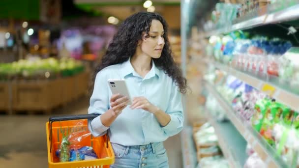 若いかなりブルネットの女性買い手顧客チェックショッピングリストを使用してスマートフォン スーパーマーケットで食料品店で購入女性買い物客ピック使用携帯電話でハイパーマーケット — ストック動画