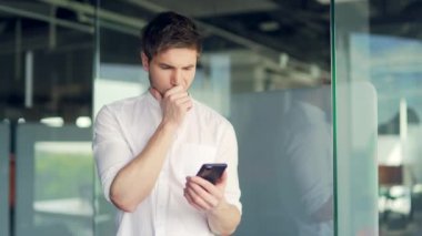 Serious thoughtful worker uses smartphone looking at screenstanding in office. Young caucasian man reading, watching content, thinking. Concentrated male look at cellphone screen browse internet