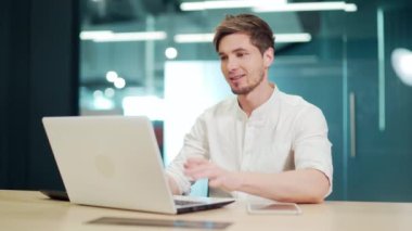 Young smiling cheerful man using video chat on laptop in office. Handsome male entrepreneur typing on a computer communicates online with a business partner or colleagues. Programmer talking remotely