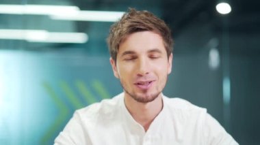 Close-up young caucasian smiling man looking at camera and talking, sitting at office desk, Webcam view of handsome guy. He talks cheerfully with the interlocutor of the virtual meeting. Head shot