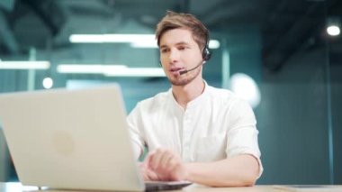 Smiling man call centre operator, customer support agent wears headset consults client online typing on laptop, talk in internet computer chat, helpline operator secretary make conference video call