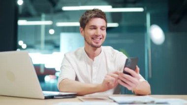 Happy employee using smartphone at work in modern office. Satisfied caucasian manager using a mobile phone while working, browsing the Internet, answering email, texting with friend or family, smiling