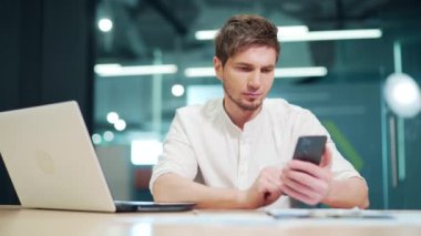 Employee using smartphone reading news at work in a modern office. Serious male checking mobile phone. Focused man tech manager using device gadget blogging typing text code at workplace indoors