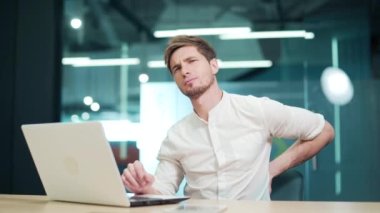 A young man feels backache while working in the office on a laptop. A male massages his back. Tired employee after long hours at work feeling discomfort in his muscles from sedentary lifestyle