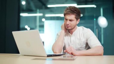 A young man feels pain in his neck while working in the office on a laptop. A male massages his neck, Feeling discomfort in the muscles from a long sedentary lifestyle. Rubs his shoulder with his hand