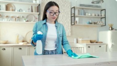 Young asian woman cleaning in the kitchen at home. A female in rubber gloves sprays furniture cleaner and wipes dust off the table with a rag. She is tidying up because she is expecting guests