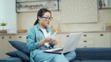 Sick with cold and runny nose, young asian woman communicates online with the help of a laptop sitting on the sofa in the living room. An upset and tired female tells about her problems to her friend